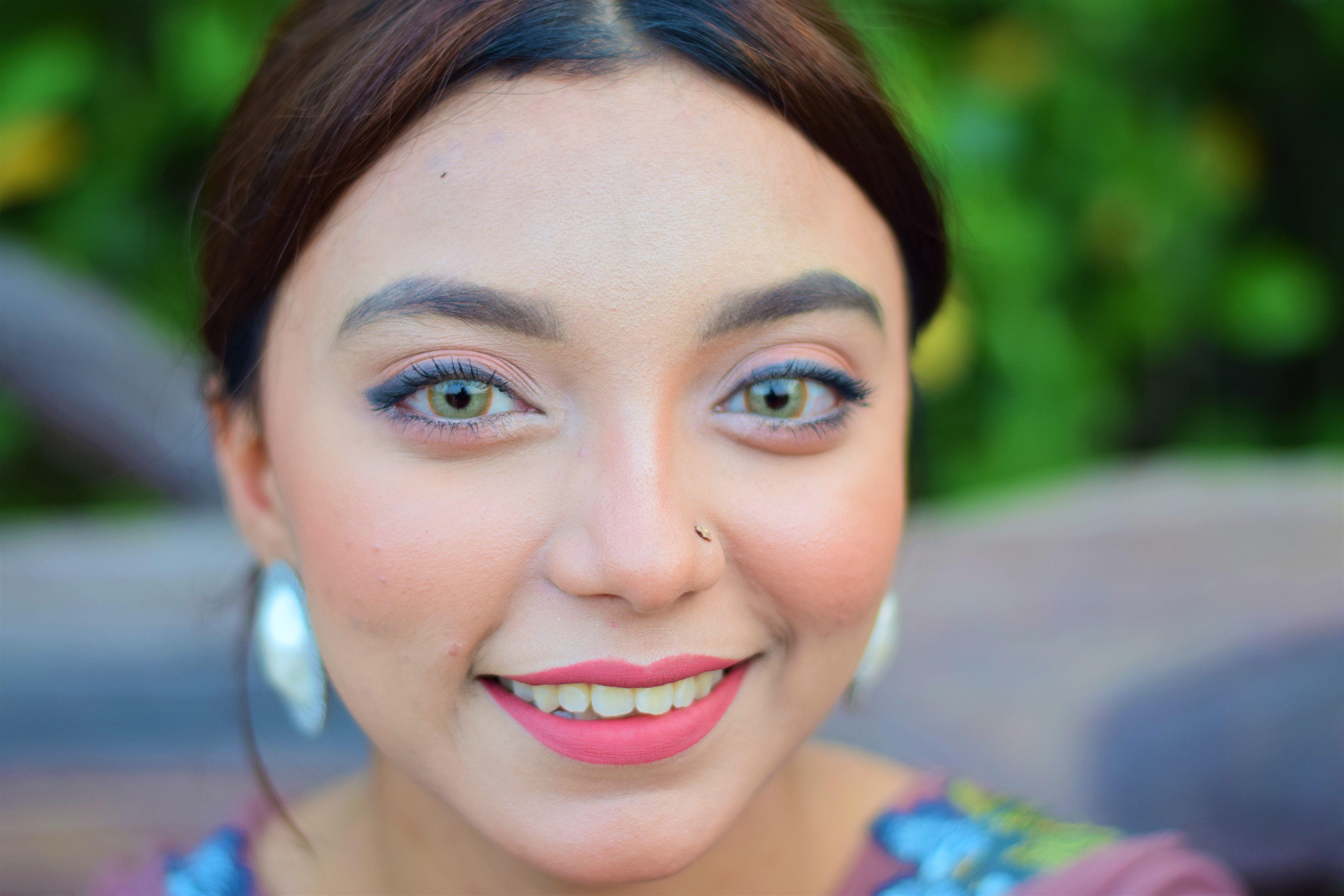 A vibrant portrait of a smiling woman with colorful eyes and nose ring, set against a green background.