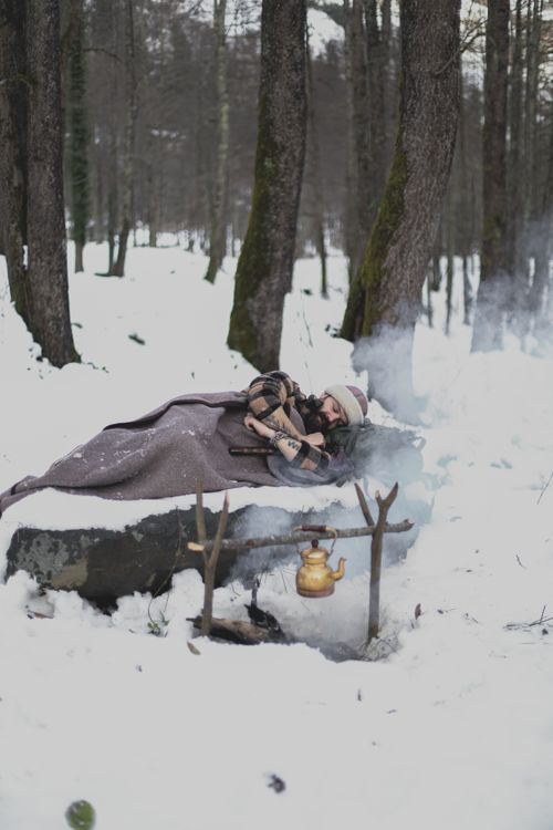 Free A person sleeps by a campfire in a snowy forest, wrapped in a blanket, with a pot hanging over the fire. Stock Photo
