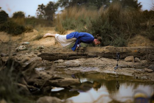 A man performs a yoga pose on a log near a pond, surrounded by a natural landscape in Dumka, India.