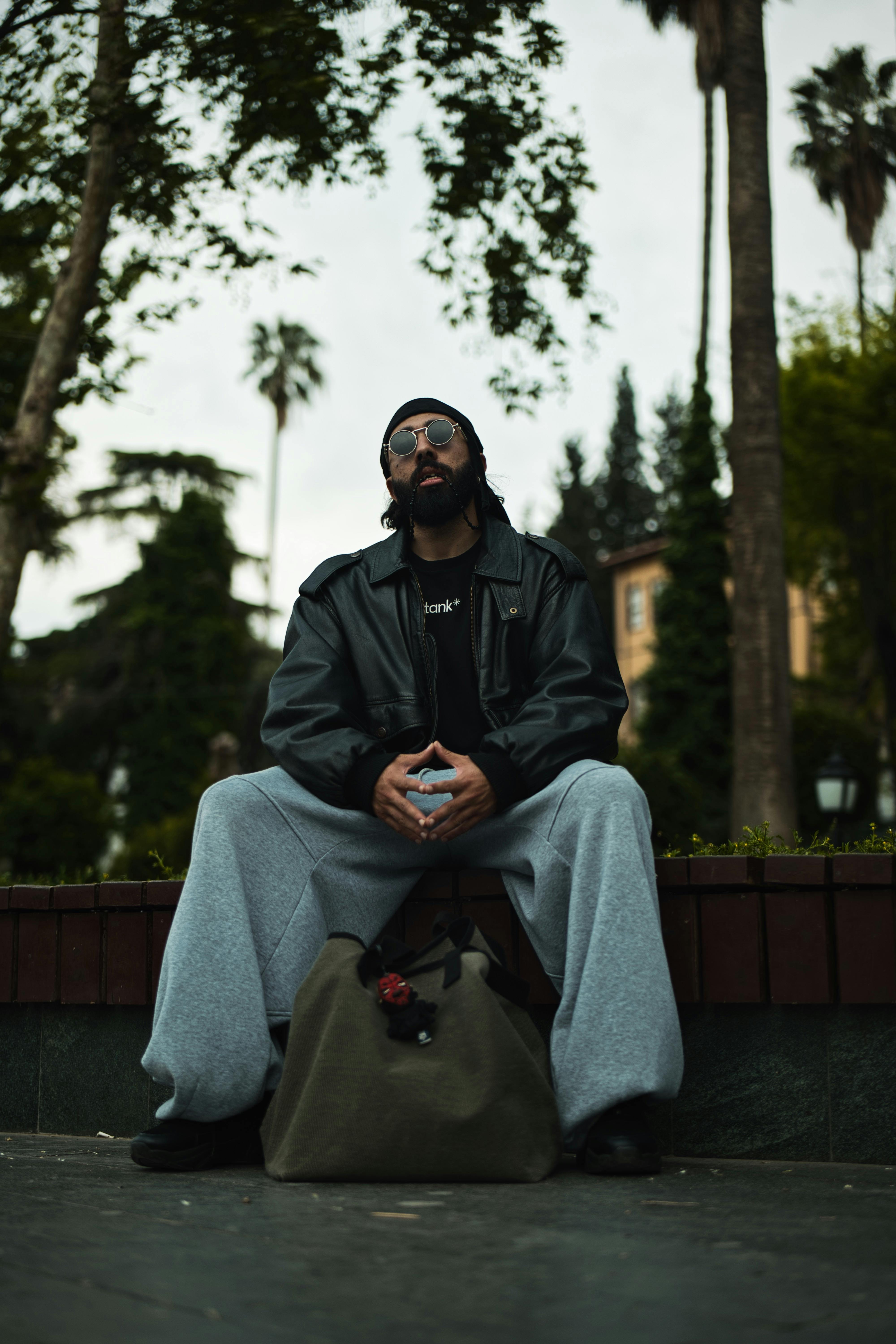 A fashionable man in sunglasses and leather jacket sitting in a city park.