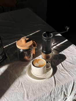 Warm sunlight on a rustic coffee setup featuring a latte with art, grinder, and jar.