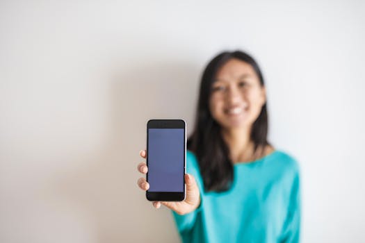 一位身穿蓝色衬衫、面带微笑的女子手持智能手机，屏幕空白，背景为白色