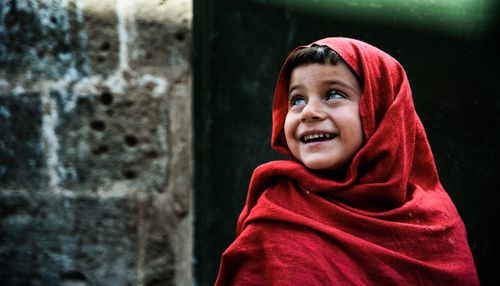 Free A joyful child wrapped in a red scarf smiles against a rustic backdrop. Stock Photo