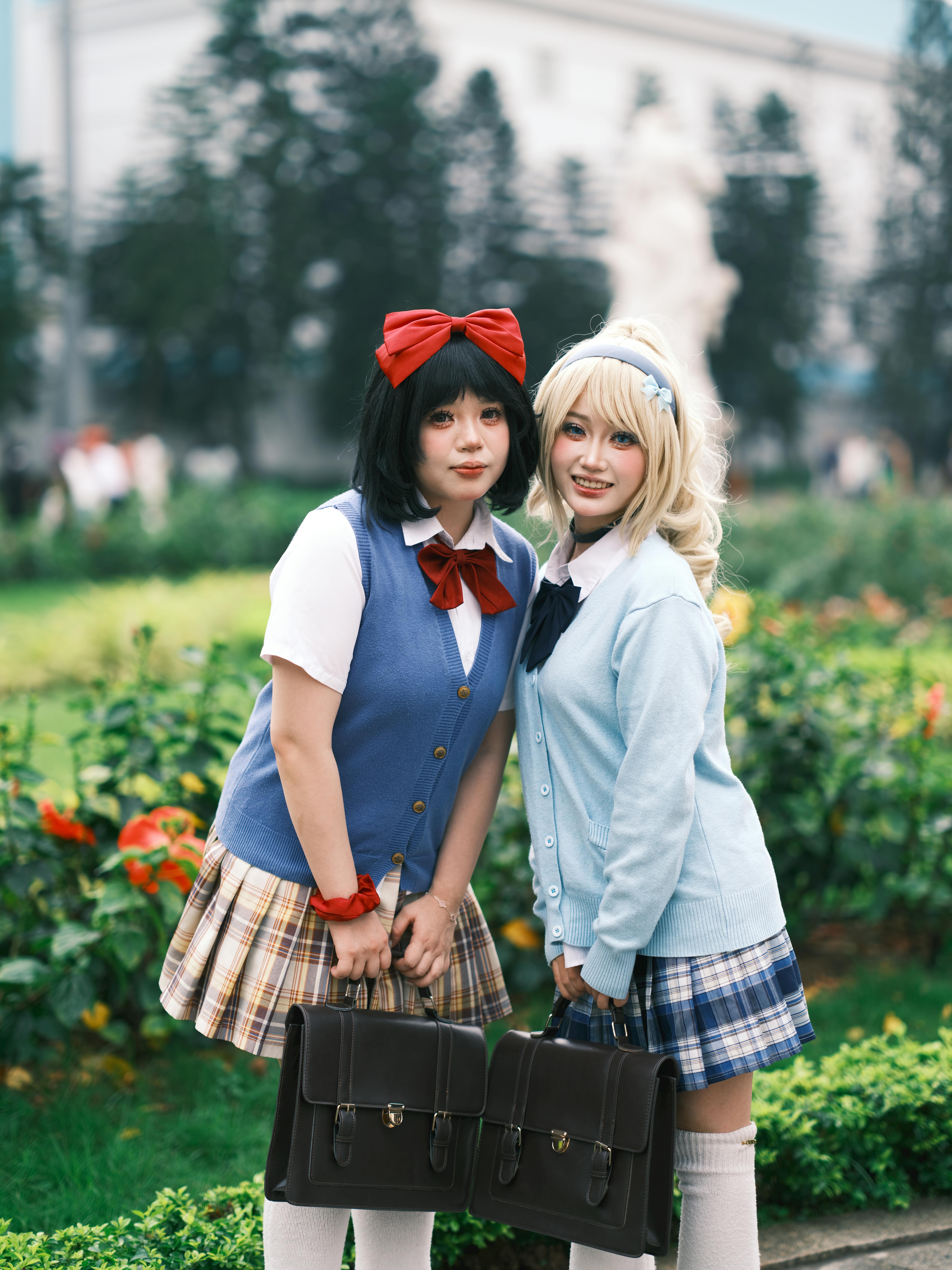 Two young women in cosplay school uniforms pose cheerfully in a garden setting.