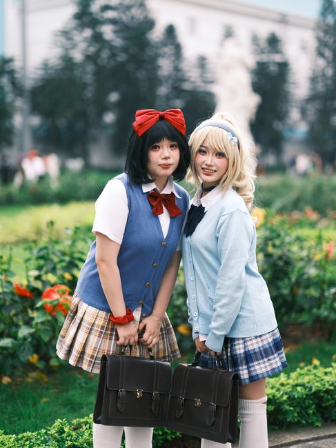 Two young women in cosplay school uniforms pose cheerfully in a garden setting.