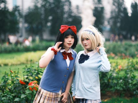 Two young women cosplay in school uniforms with vibrant bows, posing in a garden.