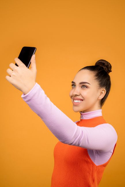 Smiling woman in casual wear taking a selfie with smartphone on vibrant orange backdrop.