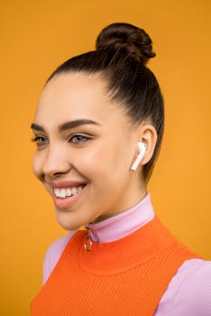 Close-up portrait of a young woman with airpods, styled in a bun against an orange background.