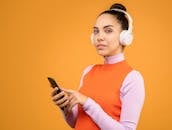 橙色背景下，一位戴着耳机、使用智能手机的时尚女性肖像