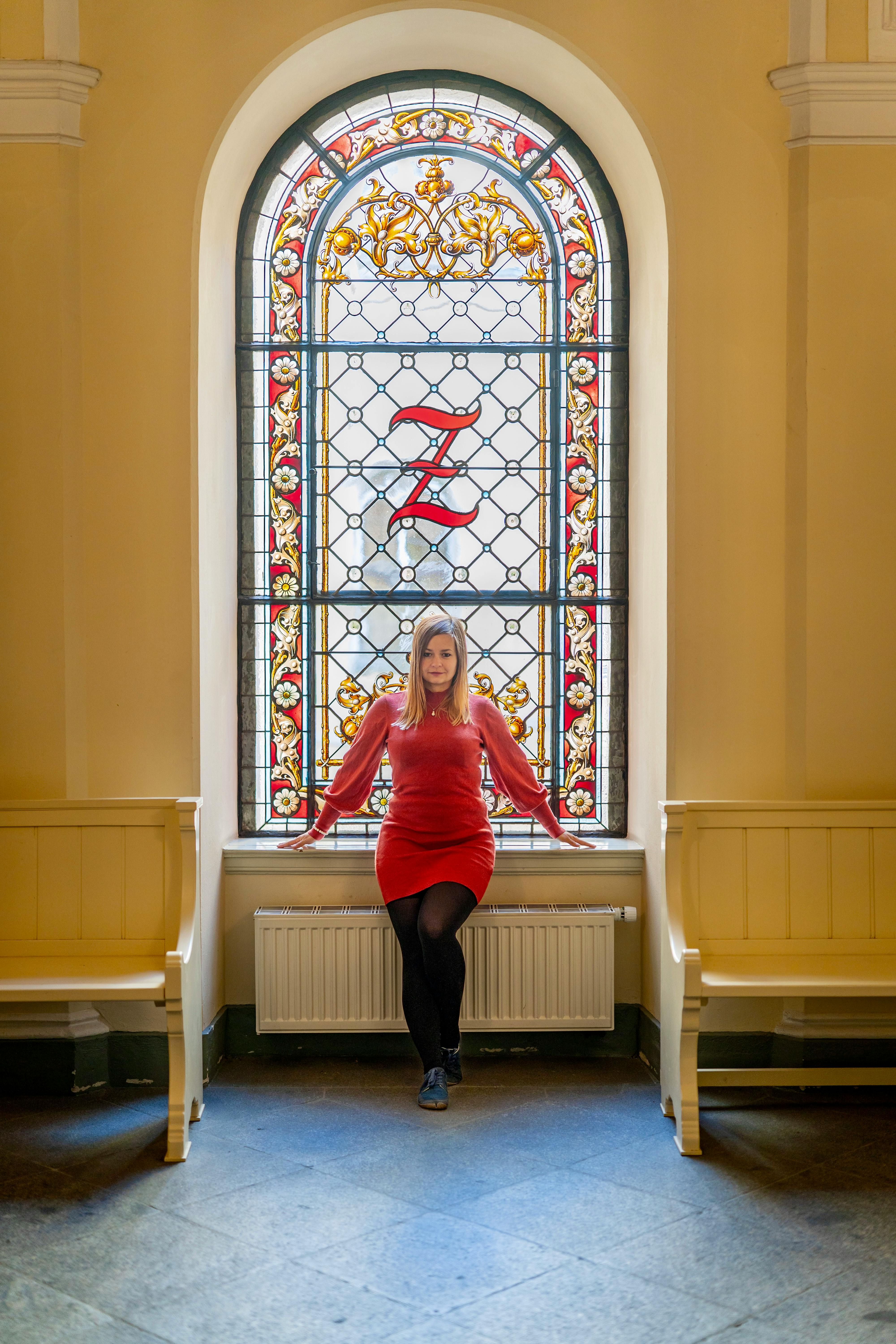 Elegant woman in red dress seated by a stained glass window in Zittau, Germany.