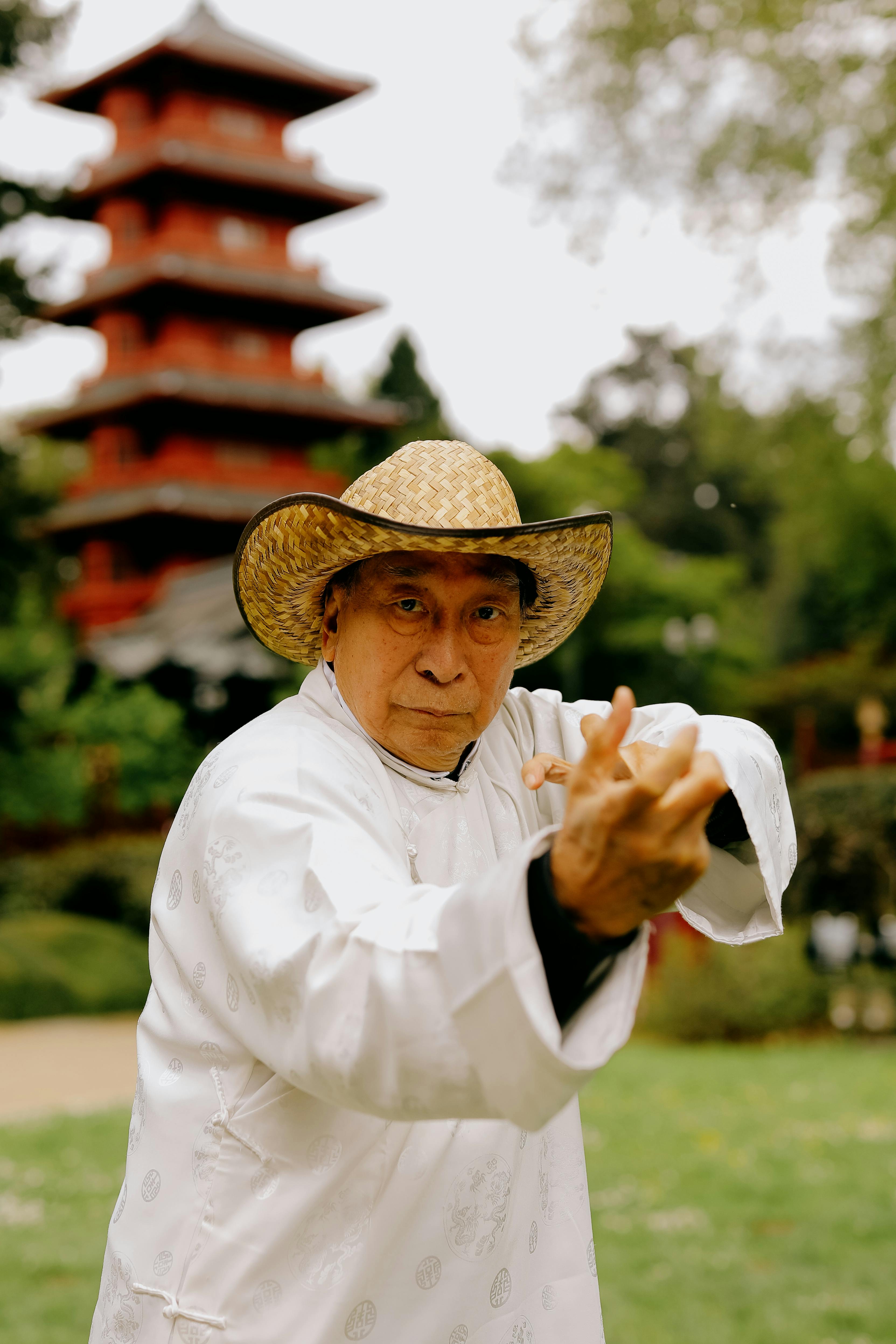 Elderly martial artist posing in front of a historic red pagoda, wearing traditional attire and a straw hat.