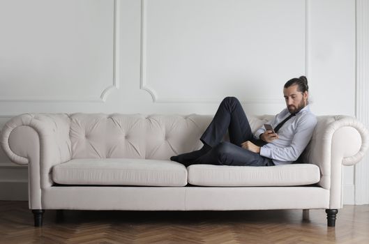 A man relaxes on a stylish white sofa in a minimalist living room, using a smartphone.