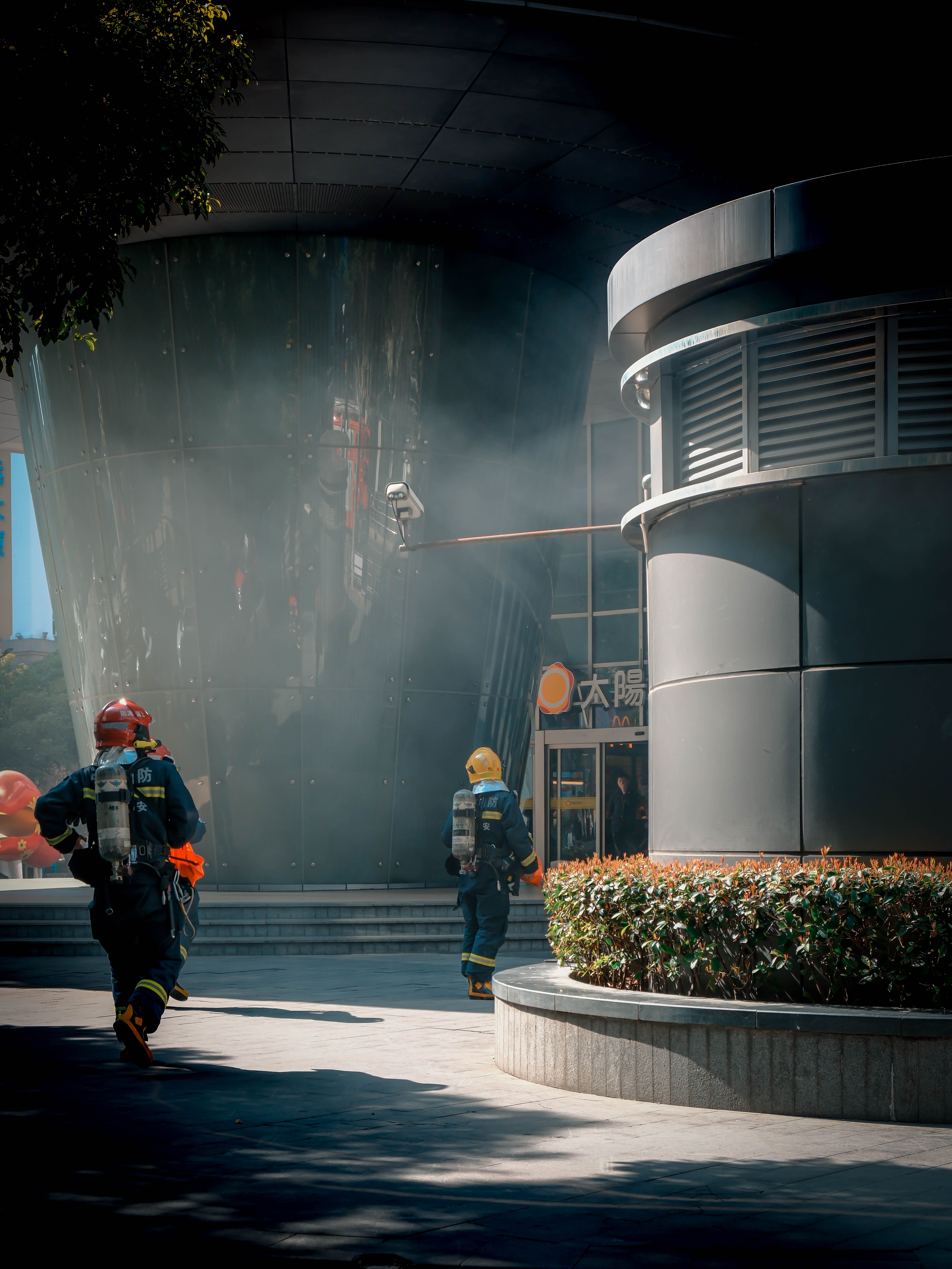 Firefighters in action outside a modern urban building, smoke visible, wearing safety gear.