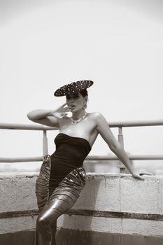 Stylish woman in glamorous attire poses outdoors with elegant hat.