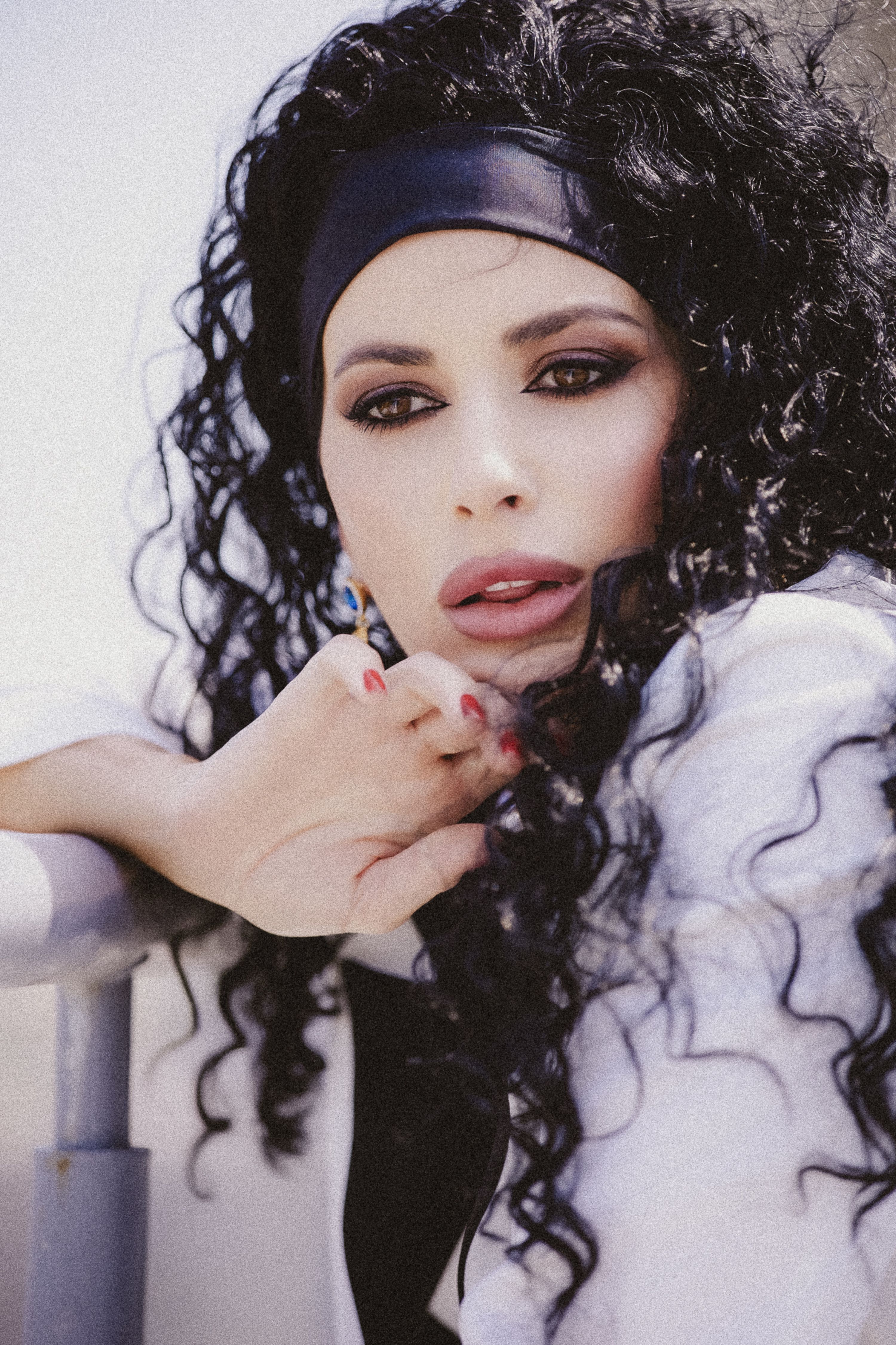 Portrait of a stylish woman with voluminous curly hair and bold makeup, leaning on a railing outdoors.