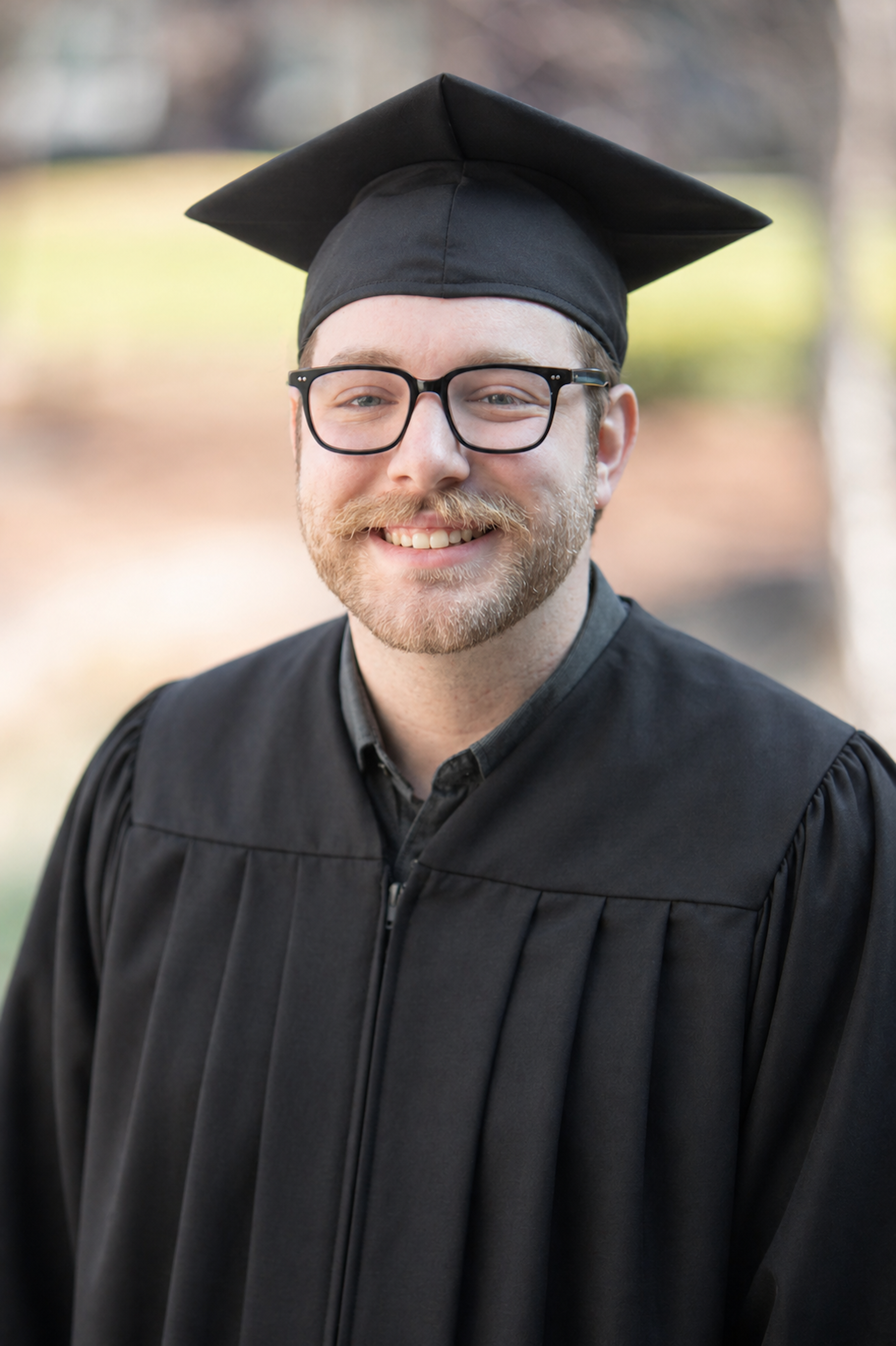 Smiling young graduate wearing cap and gown in outdoor setting.