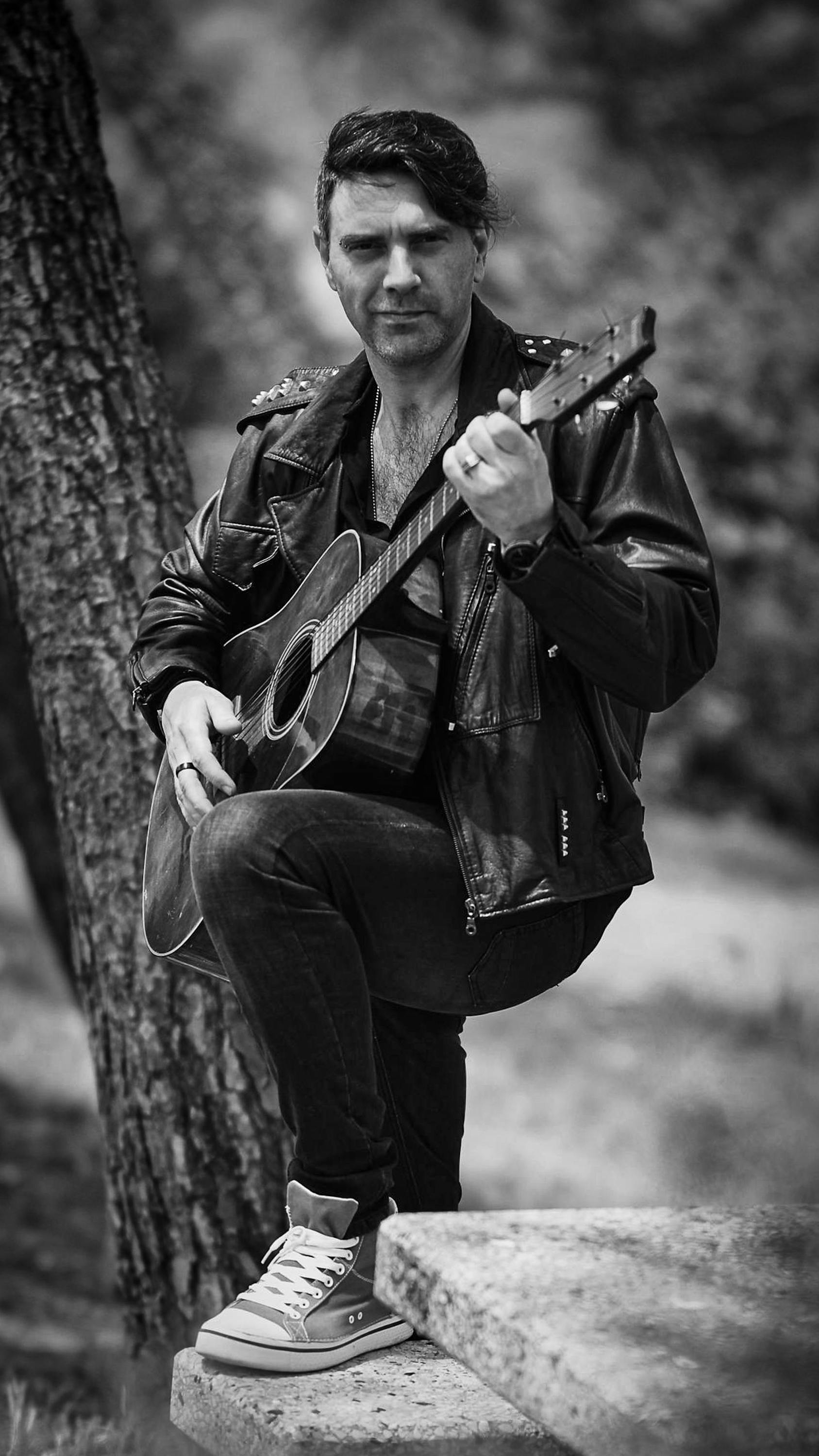 A man wearing a leather jacket plays an acoustic guitar outdoors. Black and white photo.