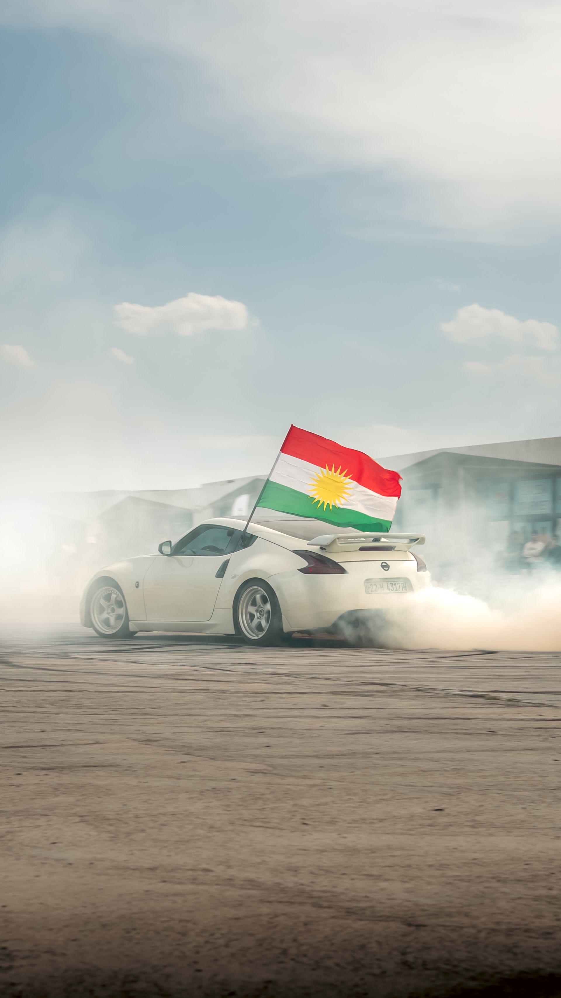 A sports car performs a drift on a sunny day, featuring a Kurdistan flag, emitting smoke.