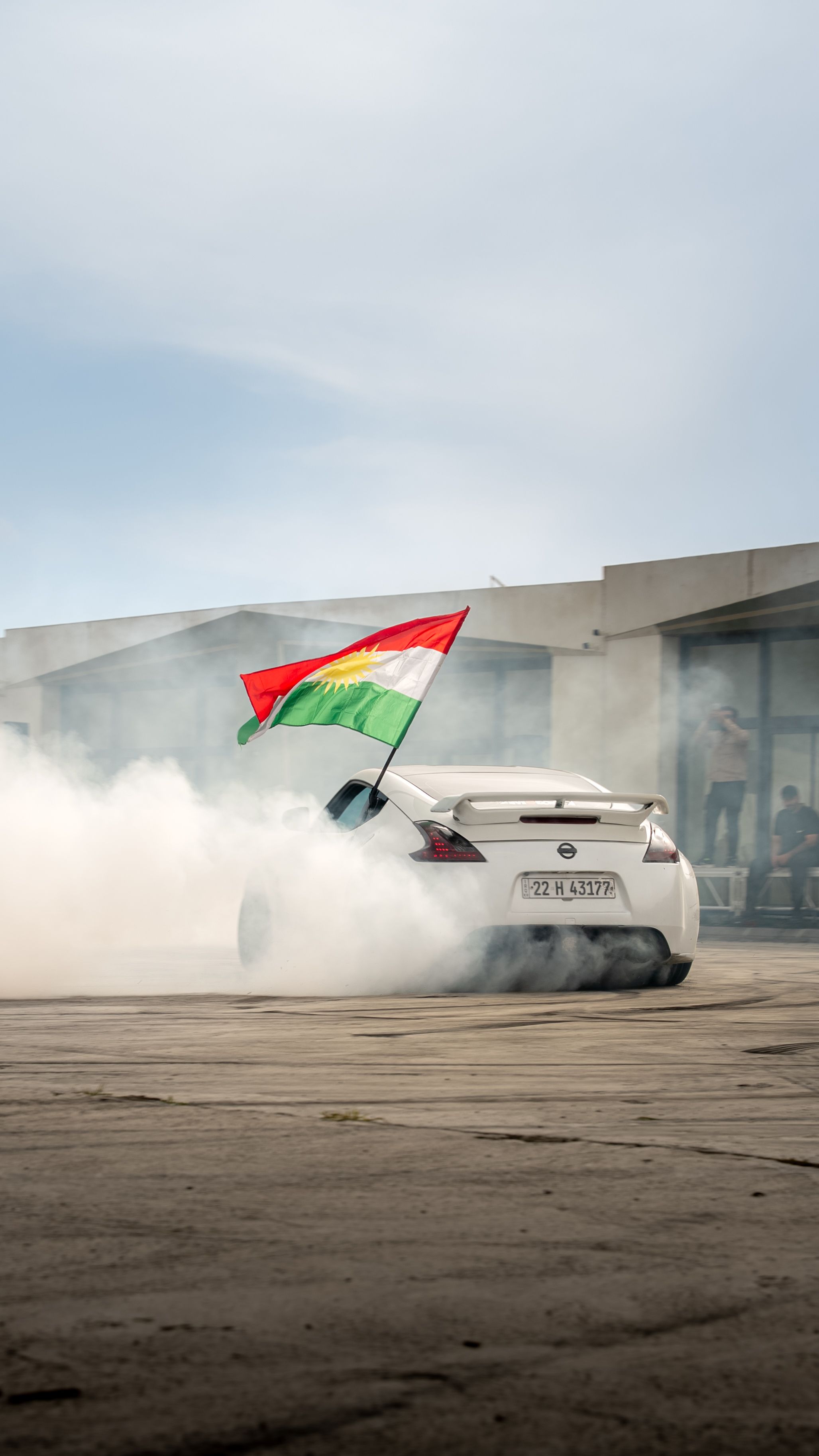 A white sports car performs a drift with smoke, displaying a national flag outdoors.