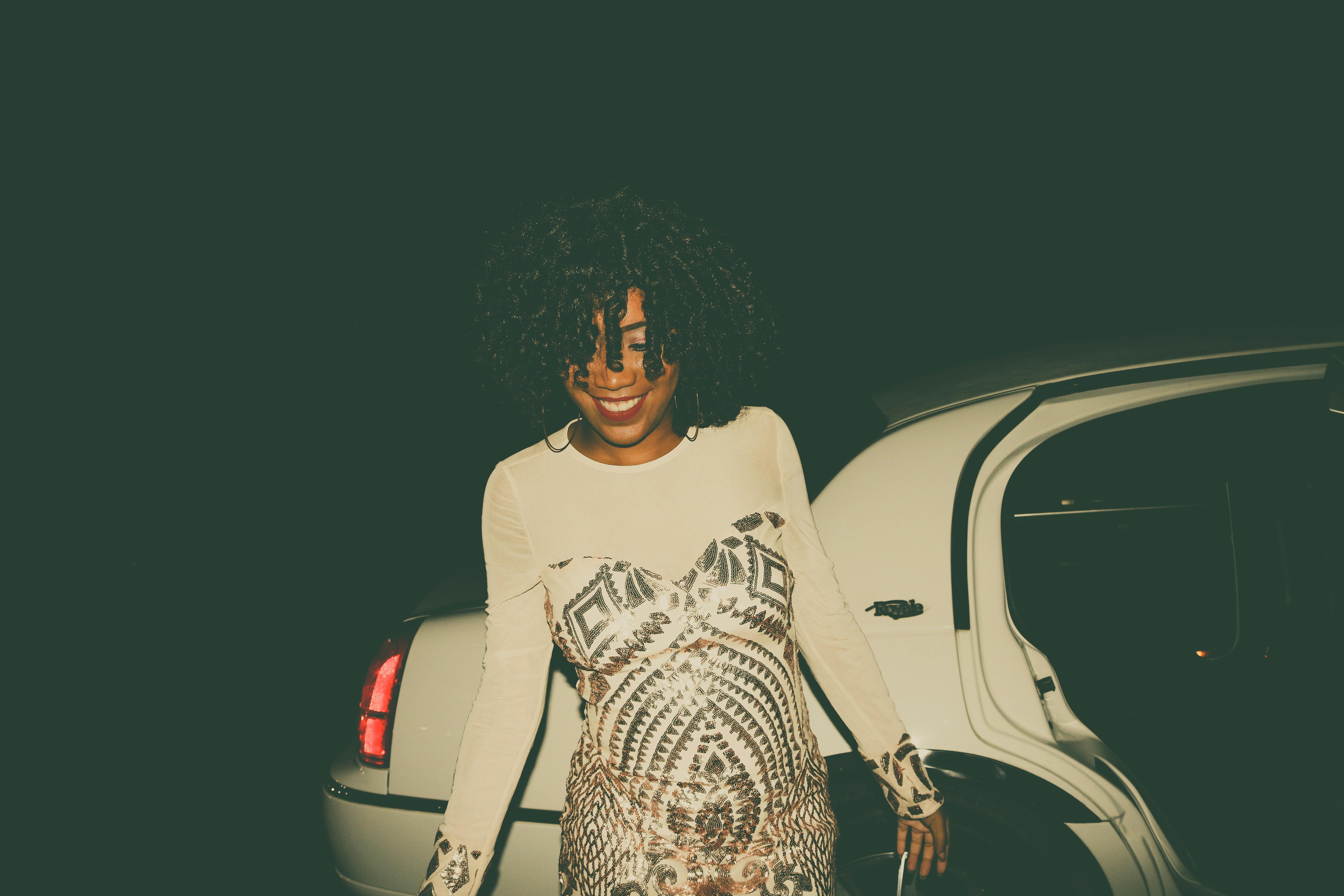 Stylish woman with curly hair smiling near a car at night, exuding confidence and elegance.