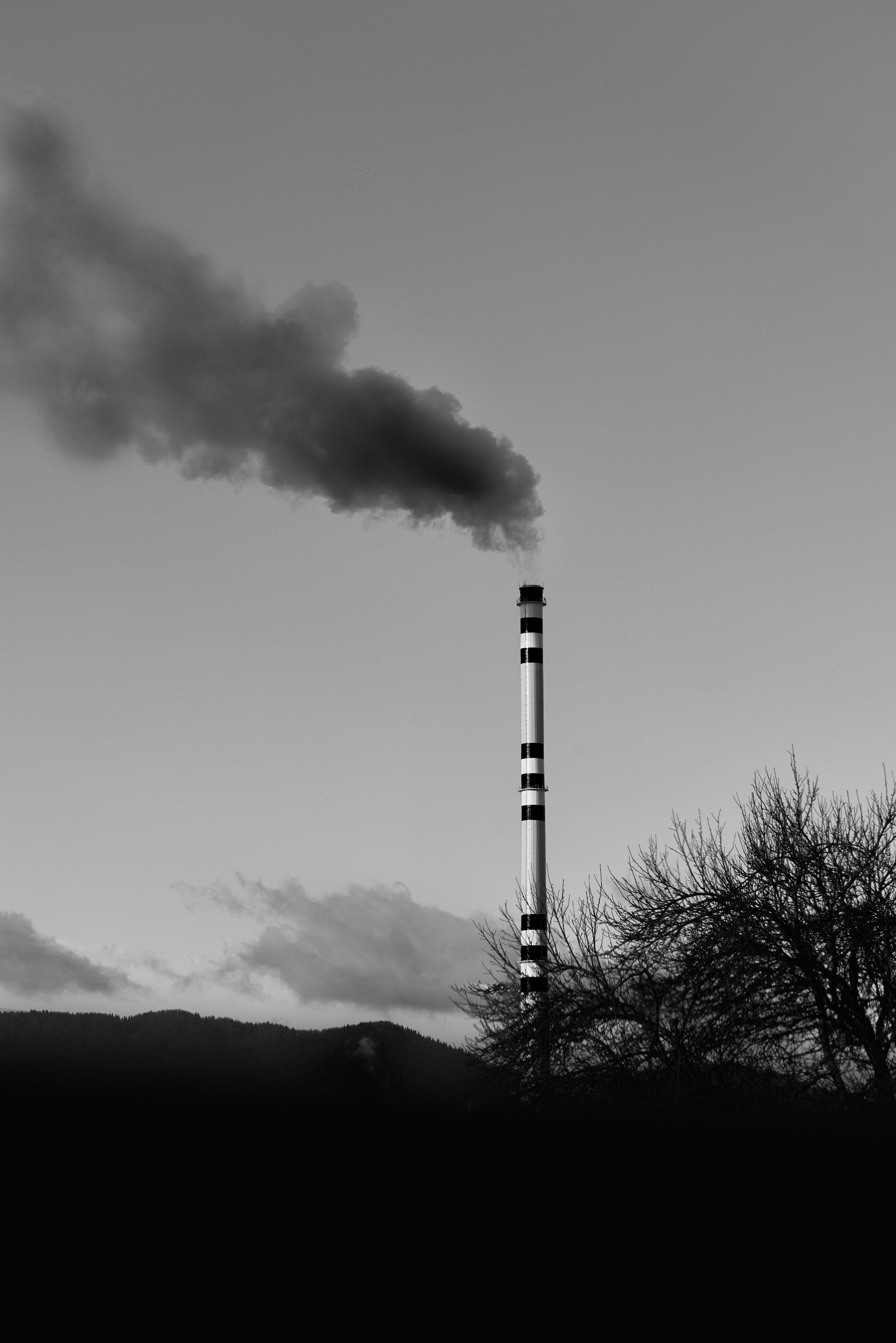 Black and white image of a smokestack releasing smoke against a cloudy sky.