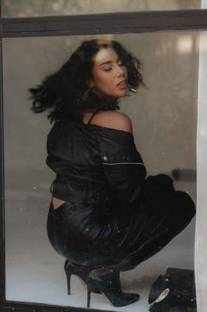 Stylish woman wearing a black leather jacket poses elegantly indoors, captured in a modern aesthetic.
