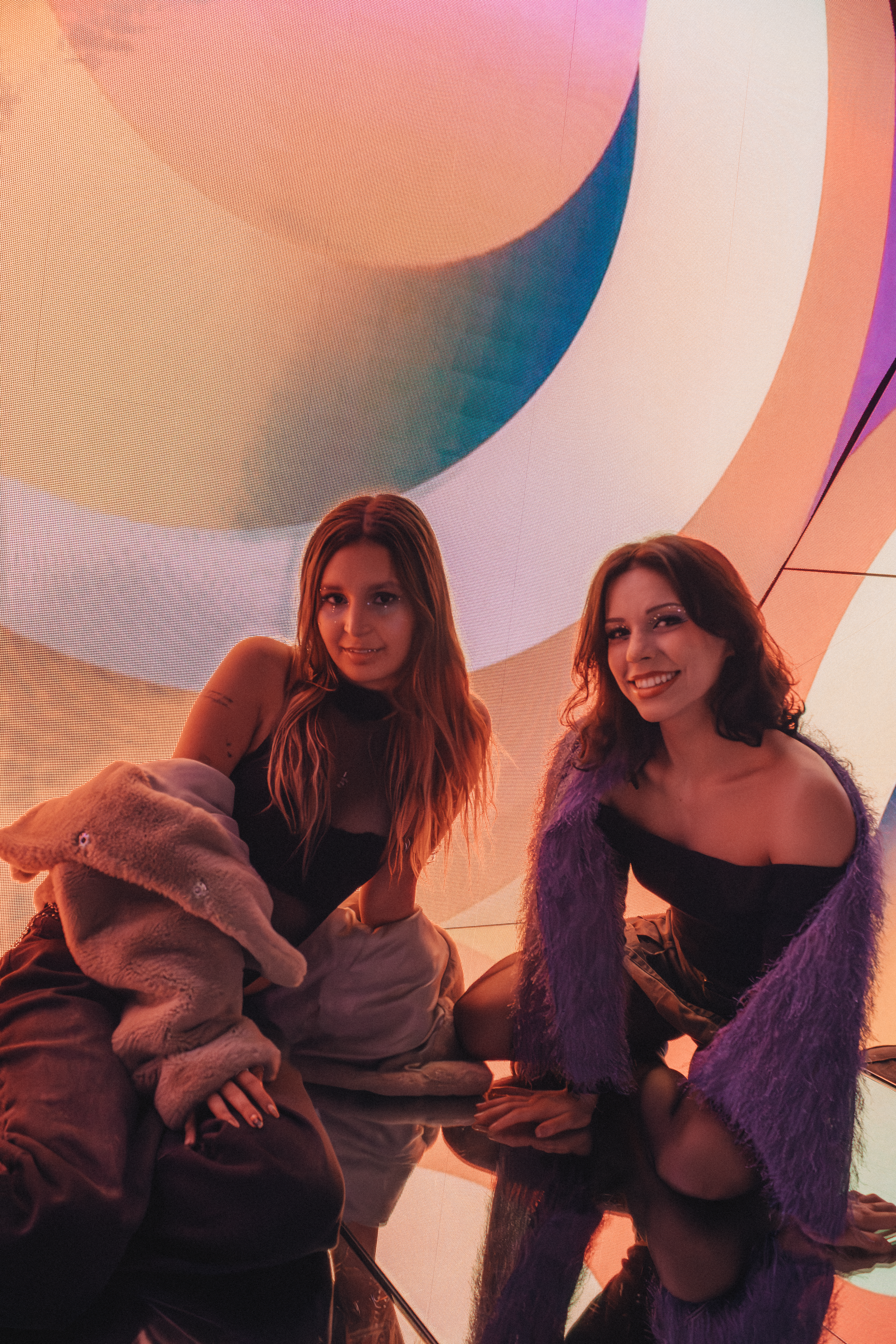 Two women in cozy outfits pose amidst vibrant abstract art indoors.