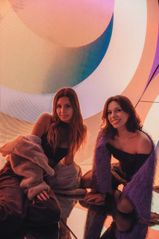 Two women in cozy outfits pose amidst vibrant abstract art indoors.