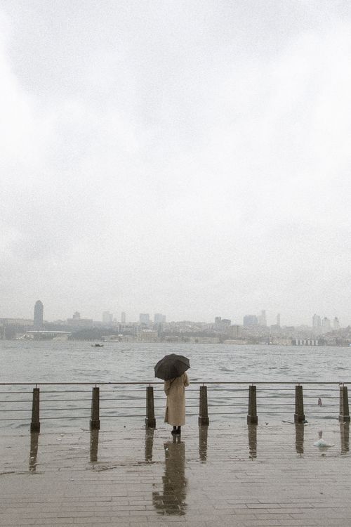 Free A lone figure stands with an umbrella by the rainy waterfront, viewing Istanbul's skyline. Stock Photo