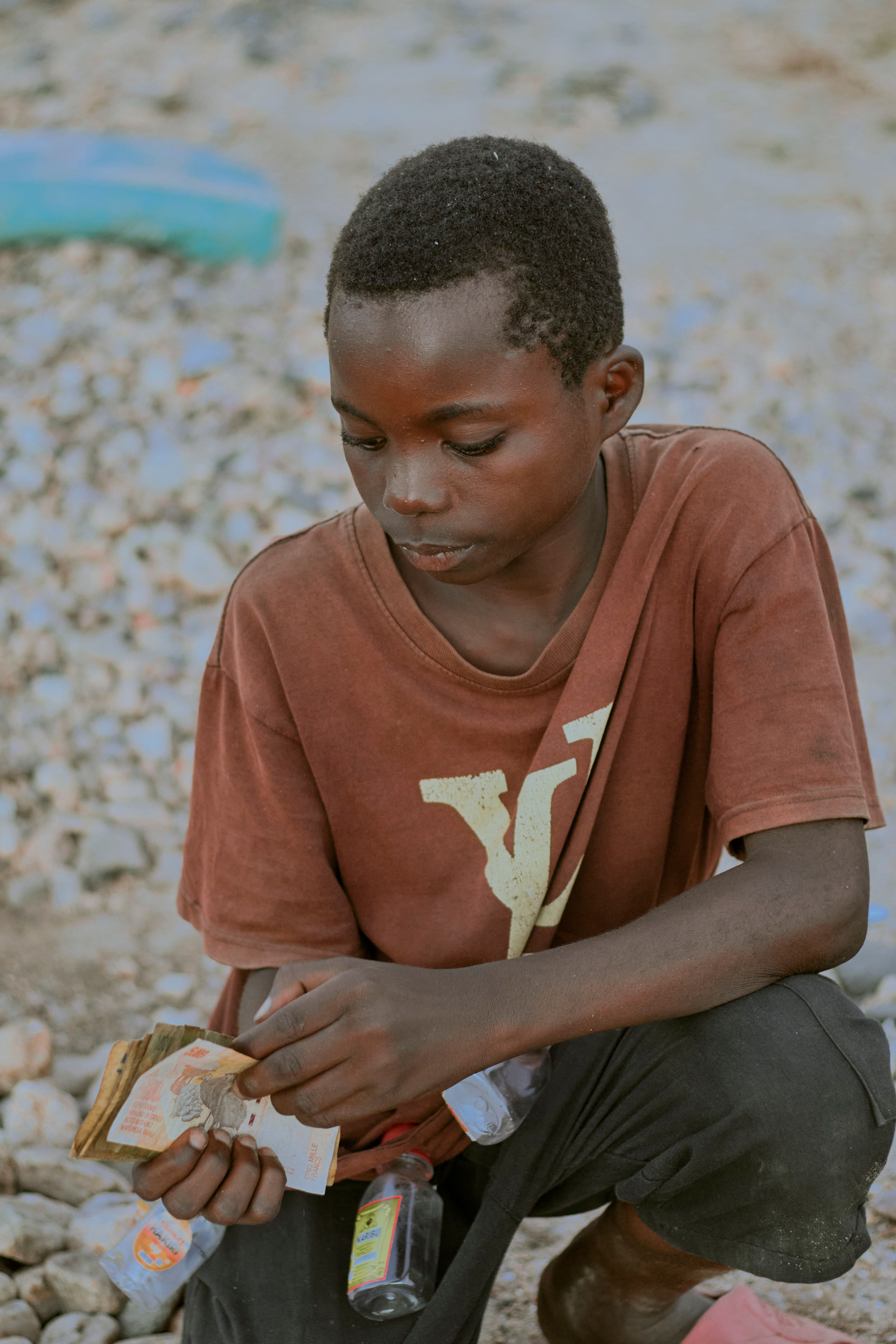 A young boy in casual clothing counts cash while seated outdoors on a rocky surface.