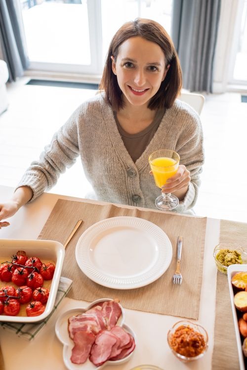 Free A cheerful woman sits at a table with fresh breakfast foods and a glass of orange juice. Stock Photo