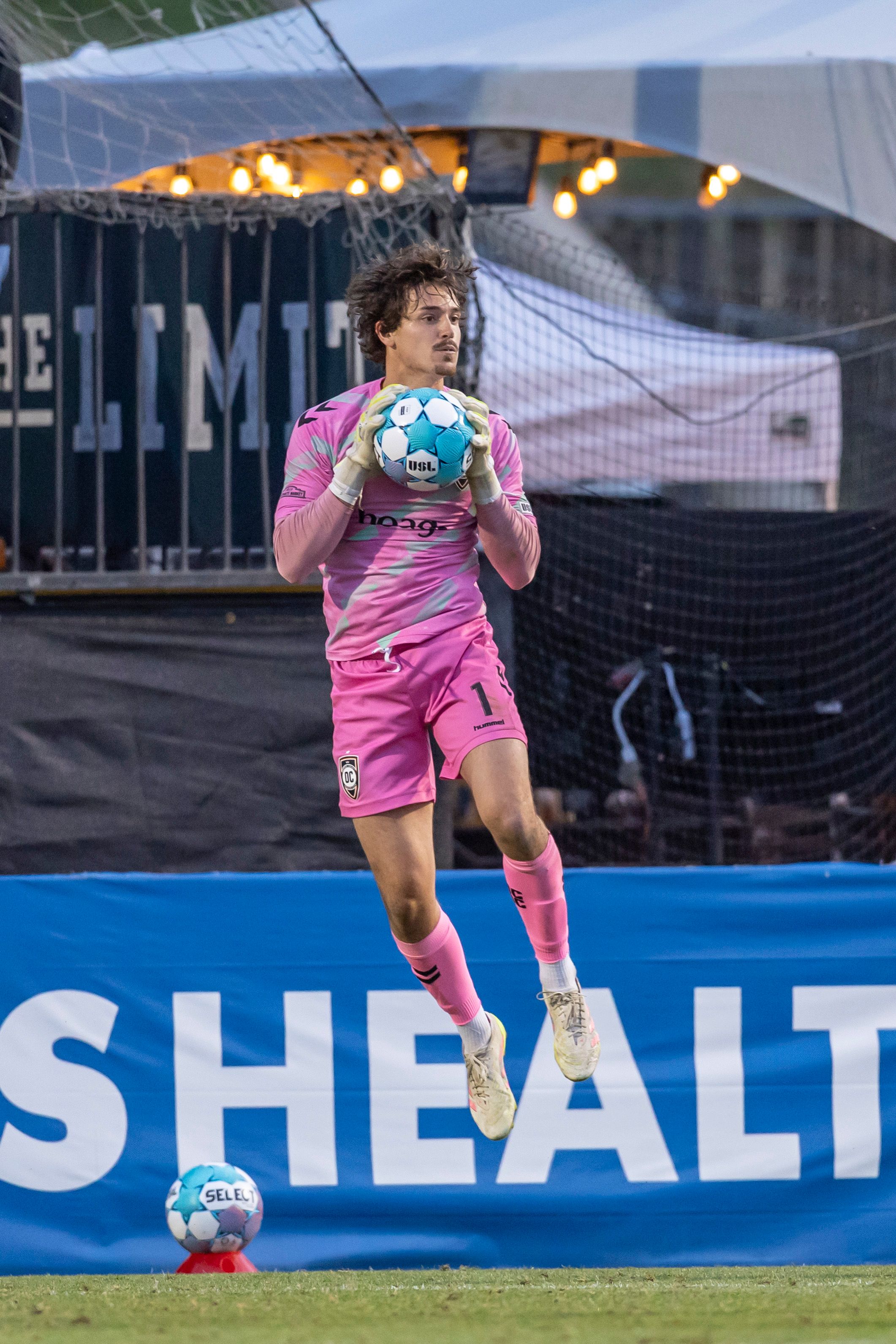 Soccer goalkeeper in pink kit jumps to catch the ball during a match in a stadium.