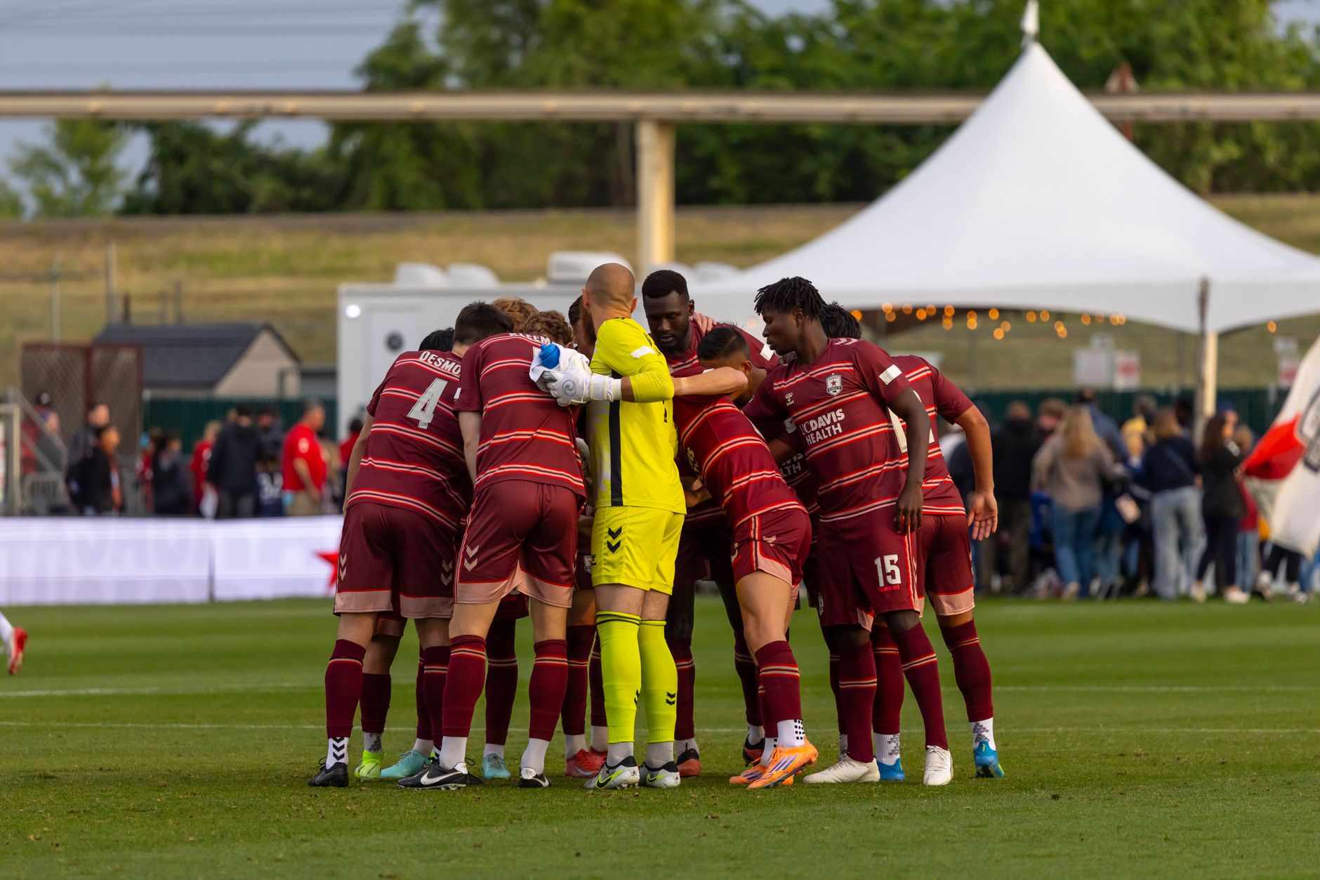 A soccer team in a huddle on the field preparing for a game outdoors on a sunny day.