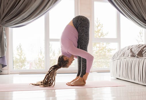 Woman performing a forward bend yoga pose in a bright, cozy room with large windows.