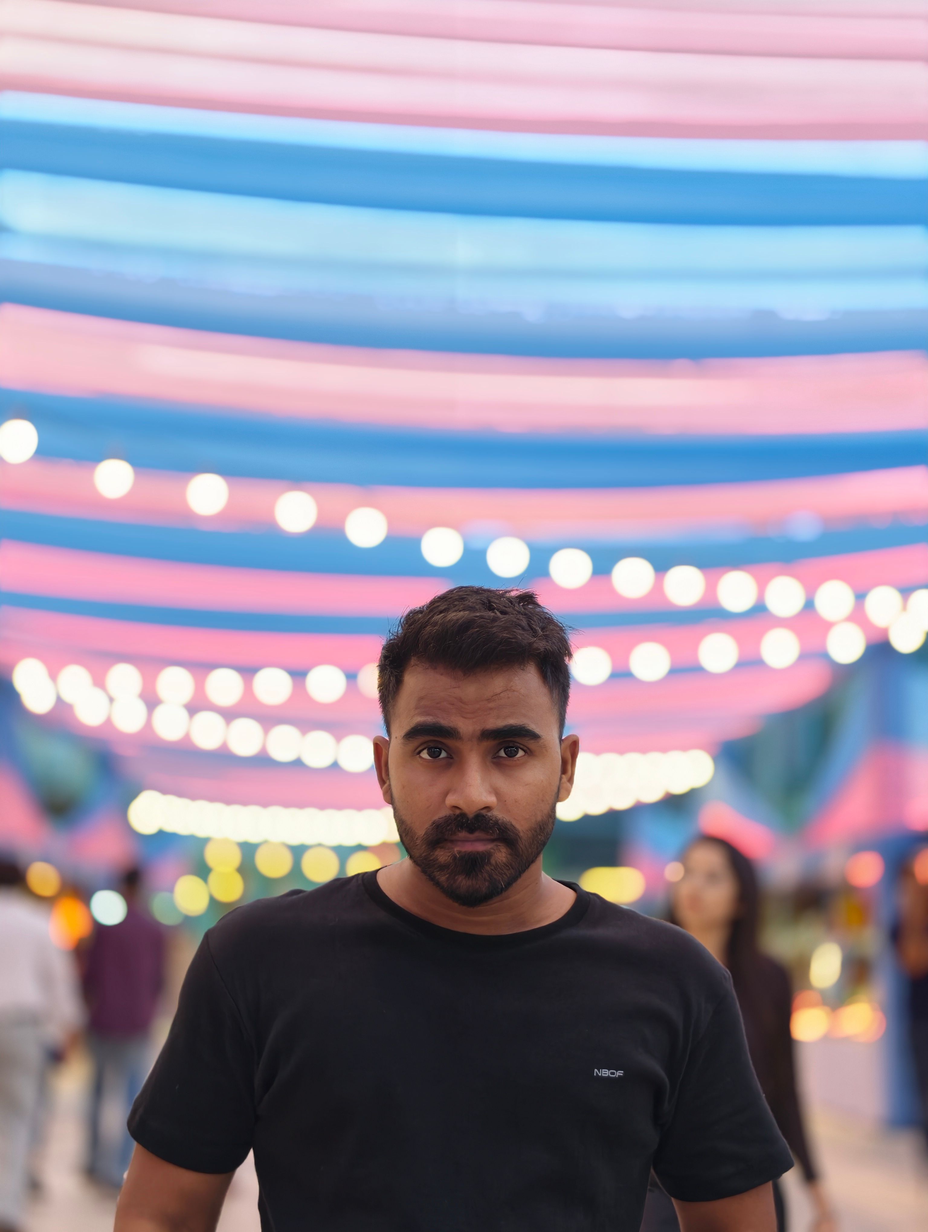 A man poses under colorful string lights at night, creating a festive atmosphere.
