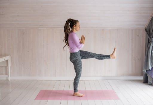 Young woman in activewear practicing a balance yoga pose on a mat indoors with natural lighting.