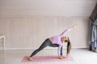 一位留着辫子的成年妇女在明亮的房间里，于粉红色垫子上表演瑜伽姿势