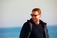 一名戴着墨镜、身穿冬季夹克的男子，在海边阳光明媚的一天中面带微笑地享受着