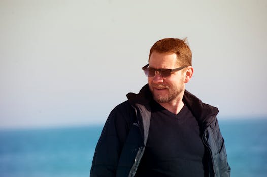 一名戴着墨镜、身穿冬季夹克的男子，在海边阳光明媚的一天中面带微笑地享受着