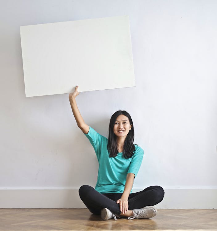 Free Cheerful Asian woman sitting cross legged on floor against white wall in empty apartment and showing white blank banner Stock Photo