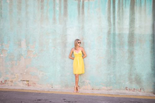 Free Fashionable woman in yellow dress and sunglasses leaning on a teal wall outdoors. Stock Photo