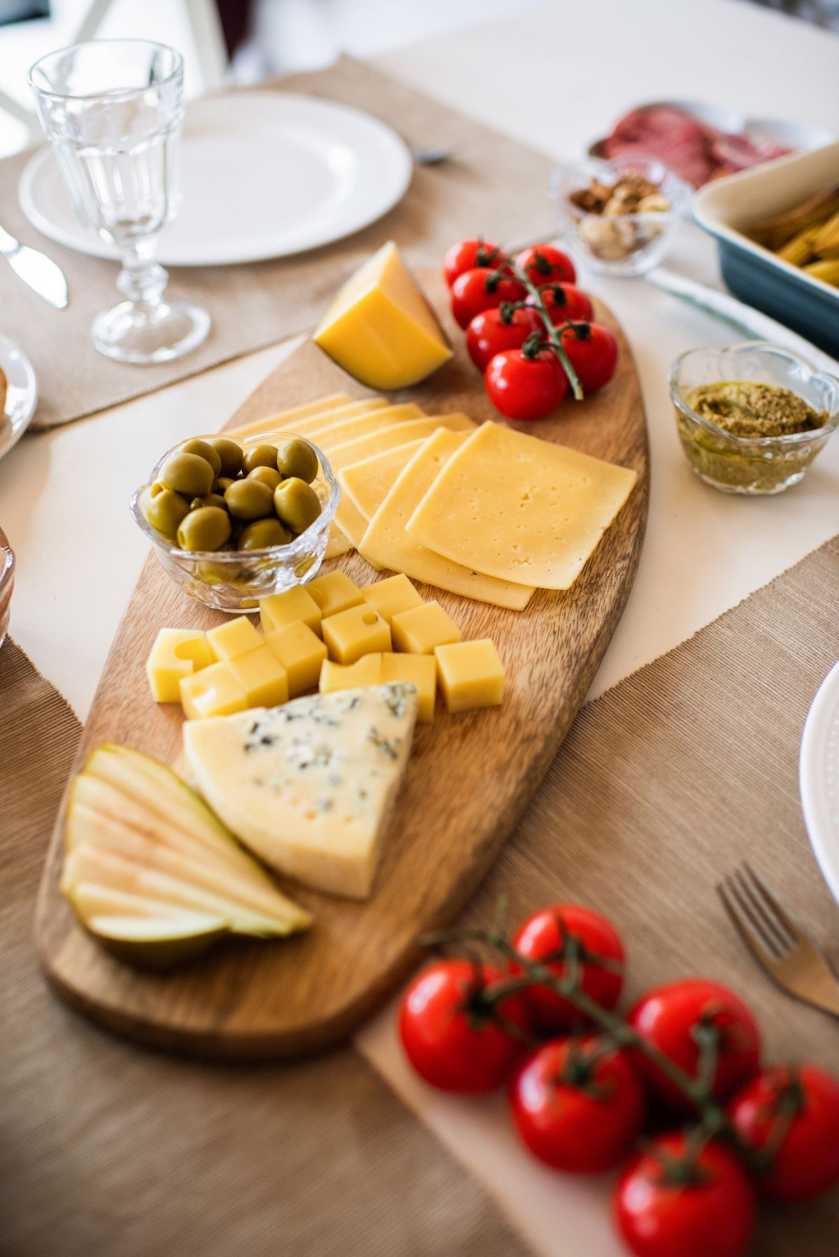 Wooden board with sliced yellow cheese, cheese cubes, blue cheese wedge, pear slices, olives and tomatoes