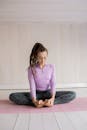 一位穿着紫色上衣和紧身裤的女子，正在室内的粉色垫子上练习坐姿瑜伽，注重放松与正念