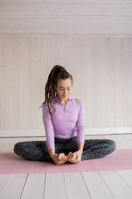Woman in purple top and leggings practicing seated yoga indoors on a pink mat, emphasizing relaxation and mindfulness.