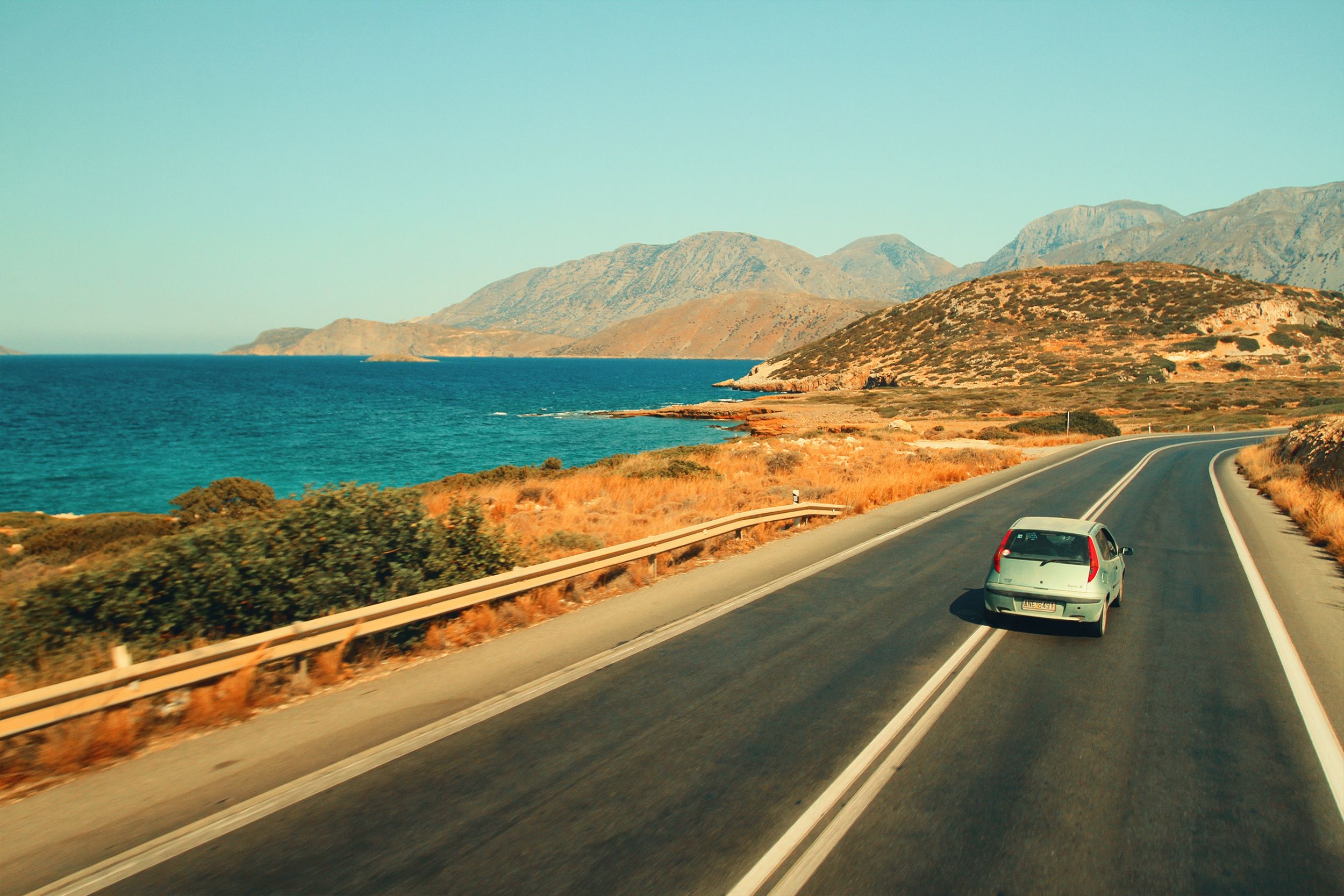 A car driving along a coastal highway with stunning ocean and mountain views under clear skies.