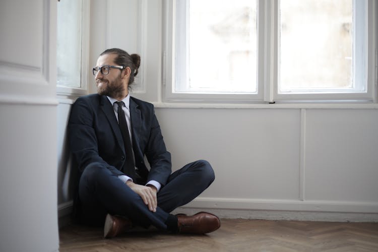 Photo Of Smiling Man In Black Suit Sitting On Brown Wooden Floor By The Corner Looking Outside A Window