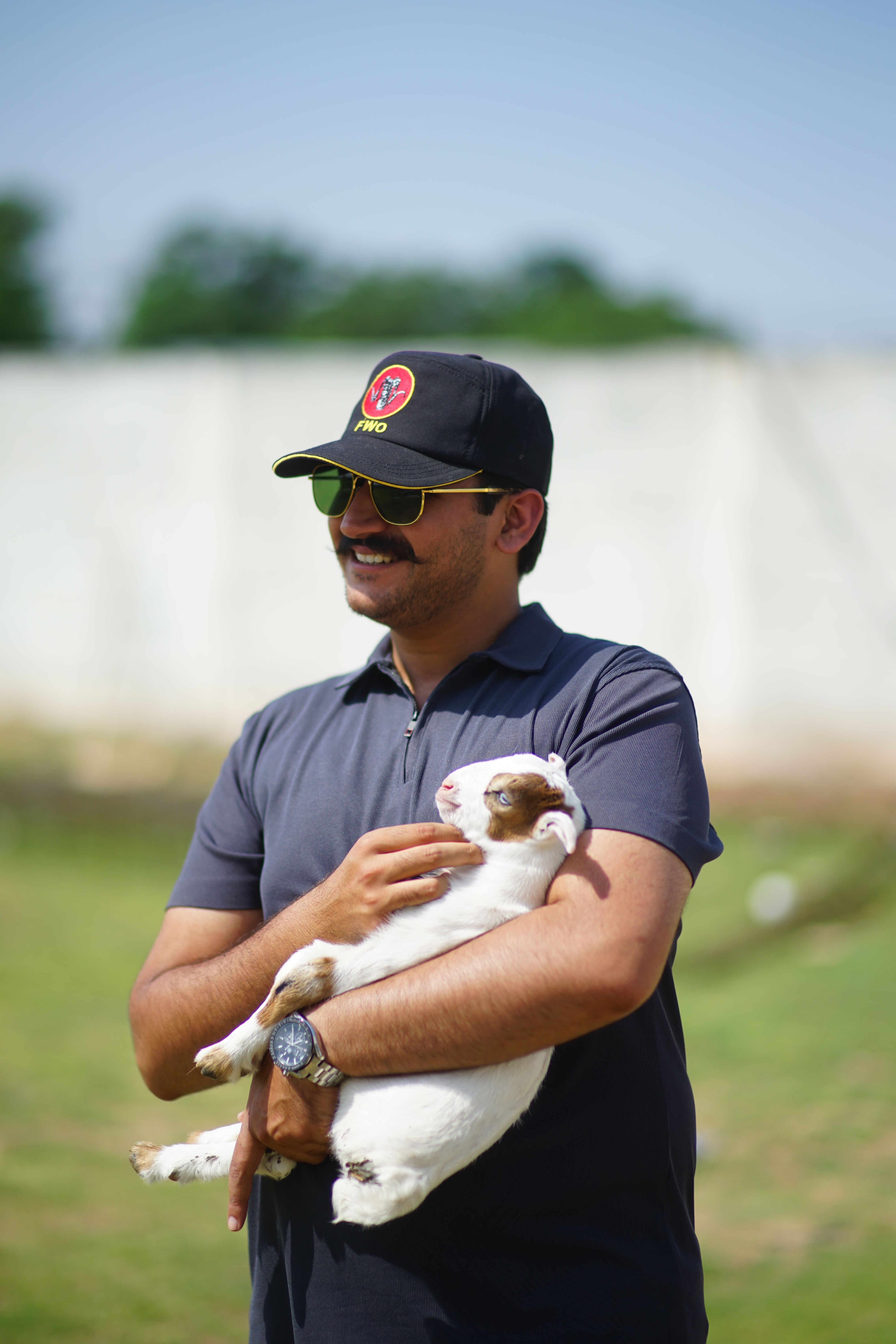 Smiling man in sunglasses holds a baby goat on a sunny day, showcasing a moment of animal companionship.