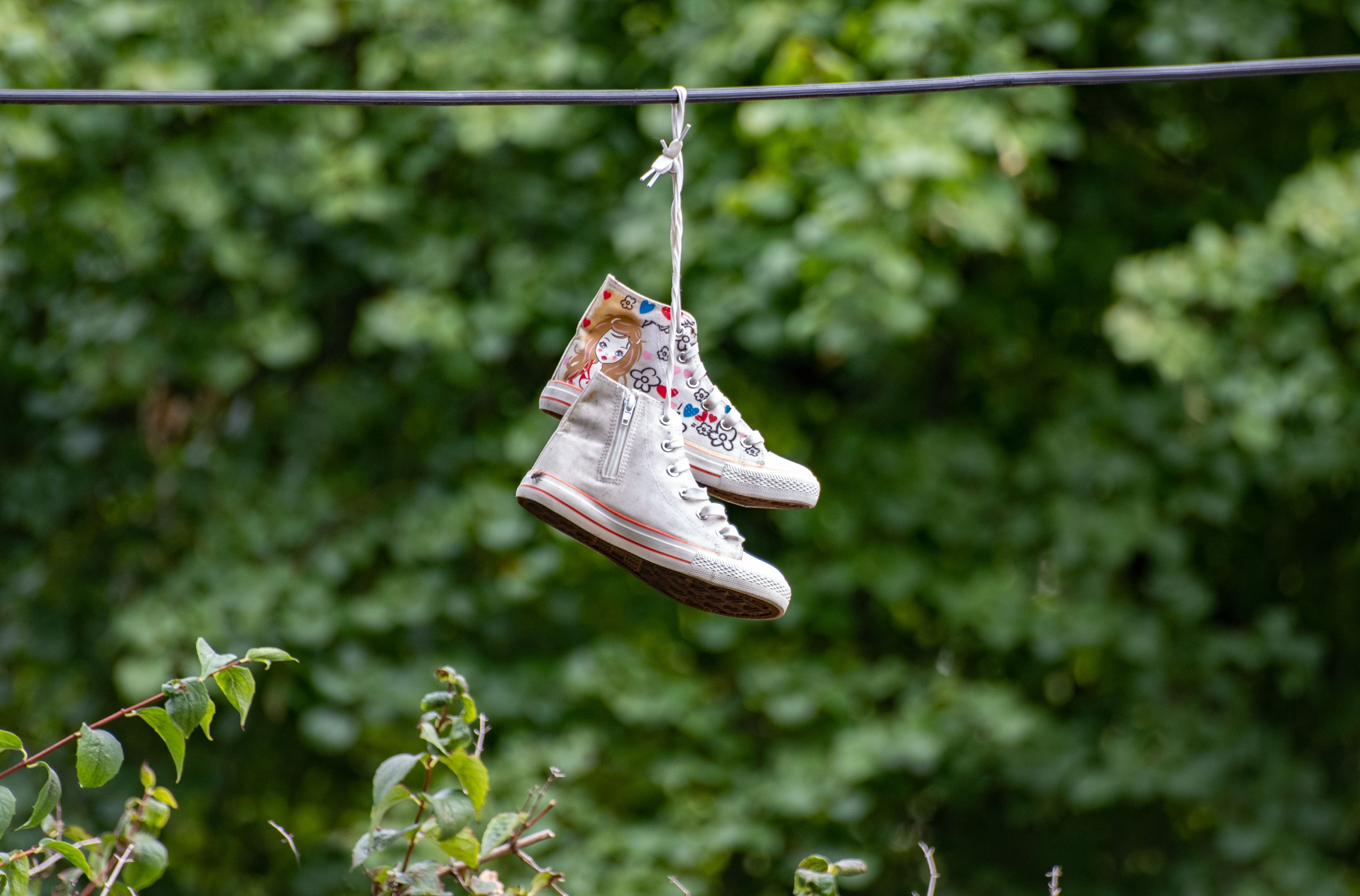 Pair of white high-top sneakers with colorful design hanging on a wire against a leafy green background.