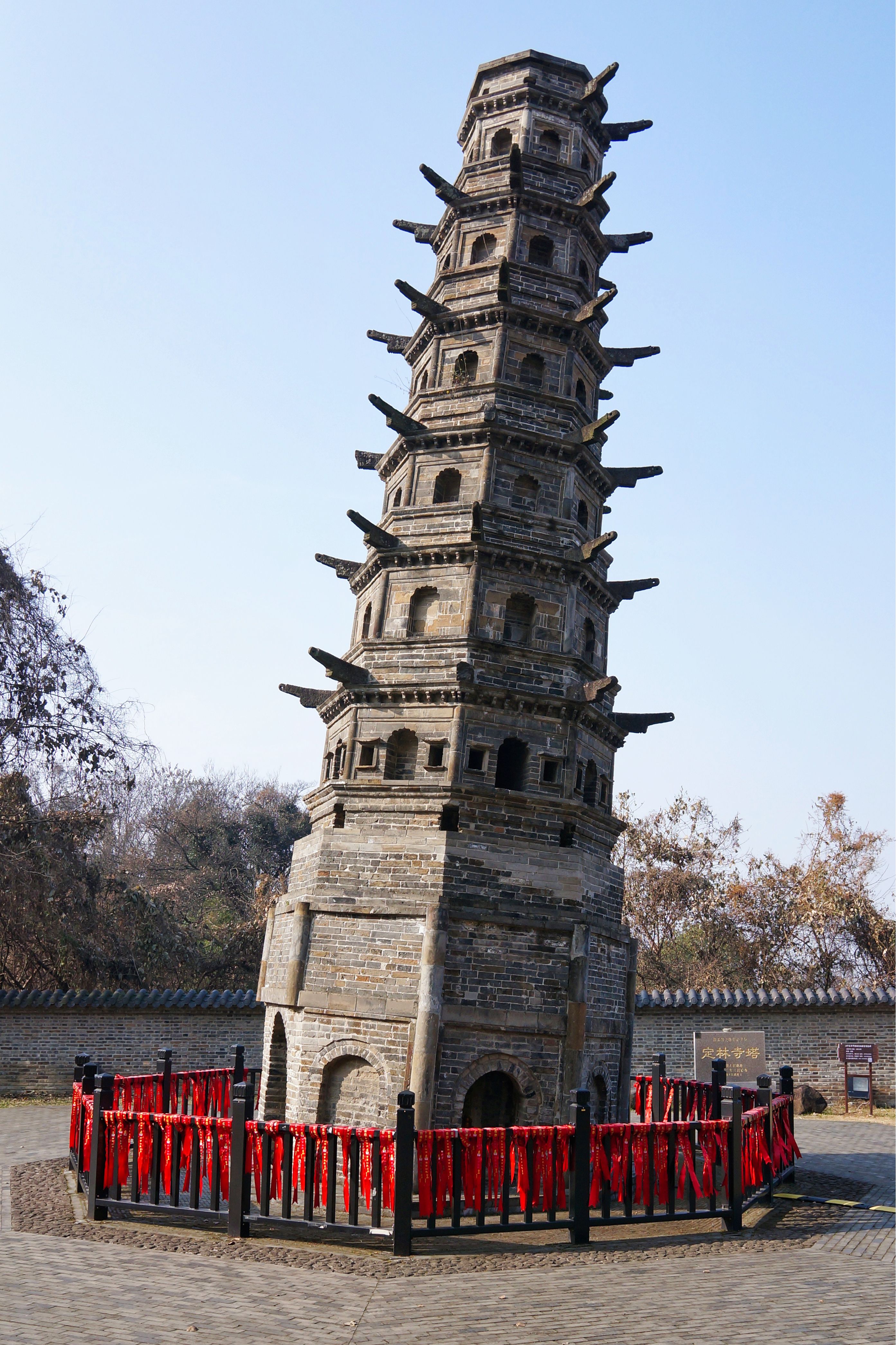 Photograph of a historic leaning brick tower in Nanjing, surrounded by red ribbons and trees.