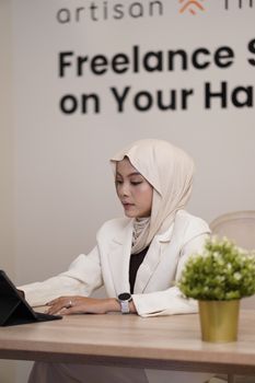 A focused woman in a white hijab and suit works on a tablet at her desk, symbolizing professionalism.